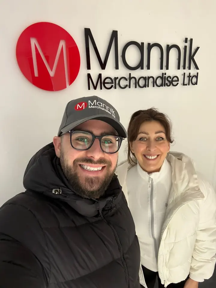 Maxx in a Mannik cap and Arlene smiling in front of the Mannik logo sign